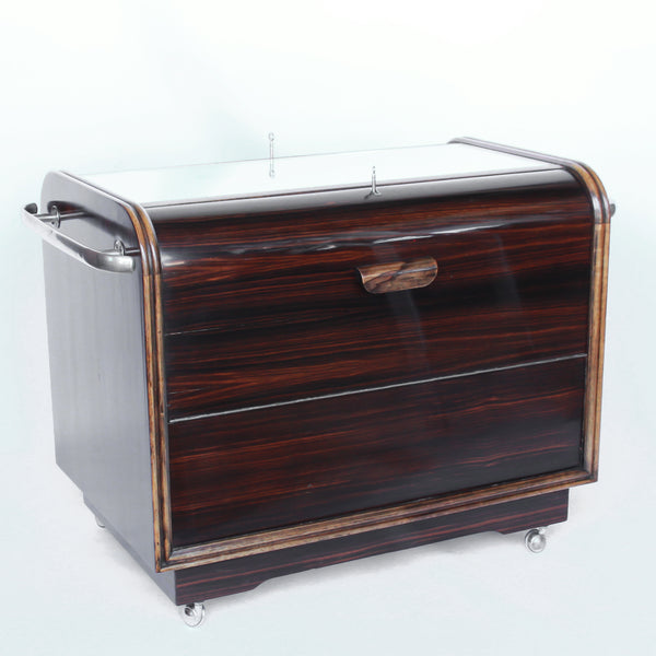 An Art Deco serving/drinks trolley with mirrored top. Double ice-box to one side with drinks cabinet to opposite side, including lower compartment for glasses. Macassar ebony throughout with walnut banding. Original chrome handle and rolling castors.