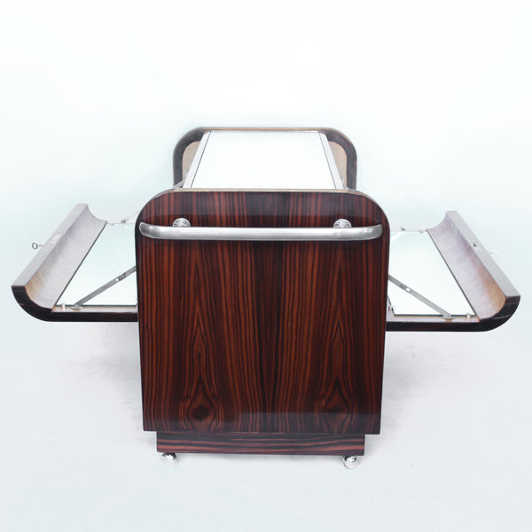 An Art Deco serving/drinks trolley with mirrored top. Double ice-box to one side with drinks cabinet to opposite side, including lower compartment for glasses. Macassar ebony throughout with walnut banding. Original chrome handle and rolling castors.